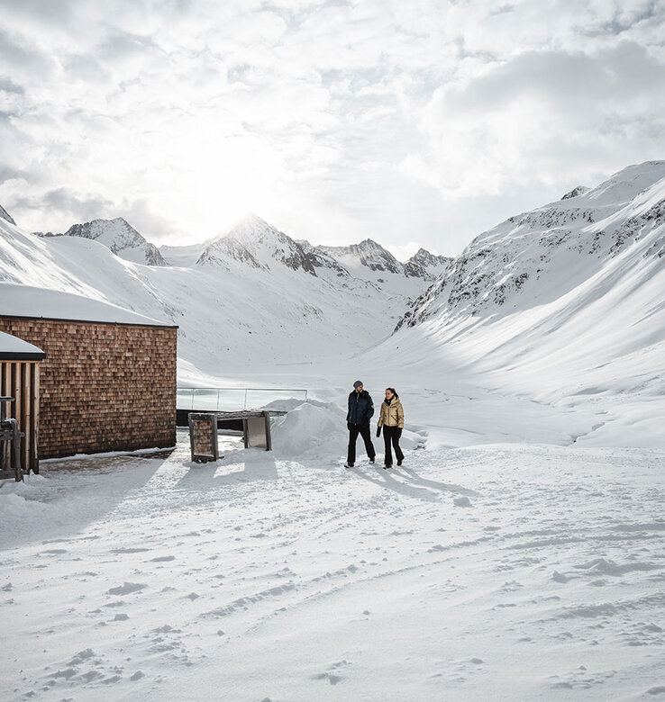Zwei Personen stehen in einer schneebedeckten Landschaft in der Nähe eines Holzgebäudes, umgeben von schneebedeckten Bergen unter einem wolkenverhangenen Himmel, durch den das Sonnenlicht durchscheint.