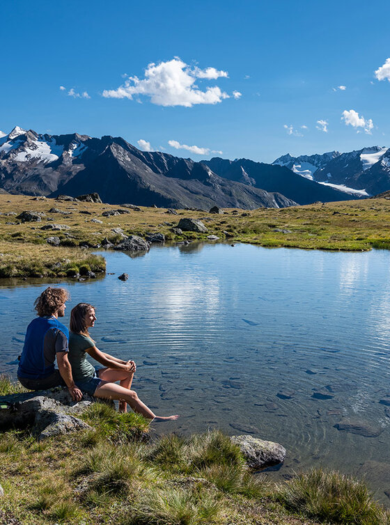 Zwei Menschen sitzen mit Rucksäcken an einem Bergsee, umgeben von Gras und schneebedeckten Gipfeln unter blauem Himmel.