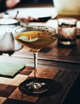 Ein Cocktail in einem Glas, garniert mit kleinen lila Blumen und einer Limettenscheibe, steht auf einem Holztisch mit einer grünen Serviette und einem verschwommenen Getränk im Hintergrund.