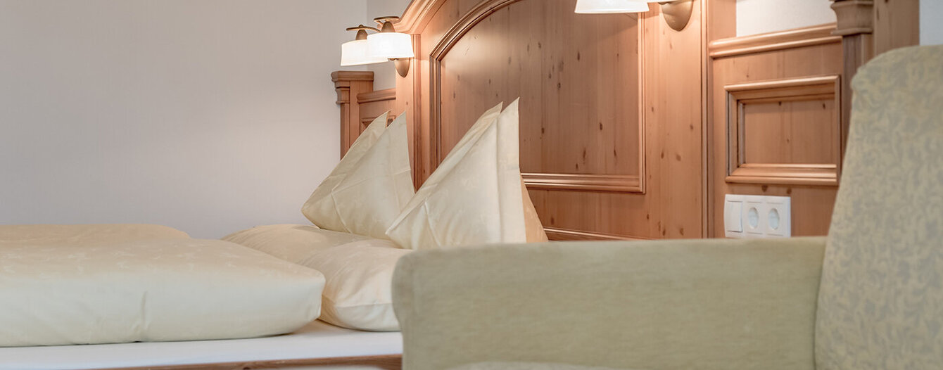 Ein gemütliches Apartmentzimmre mit einem Kopfteil aus Holz, zwei cremefarbenen Kissen, weicher Bettwäsche, eingebauten Leselampen und einem Teil eines hellbeigen Sessels im Vordergrund.