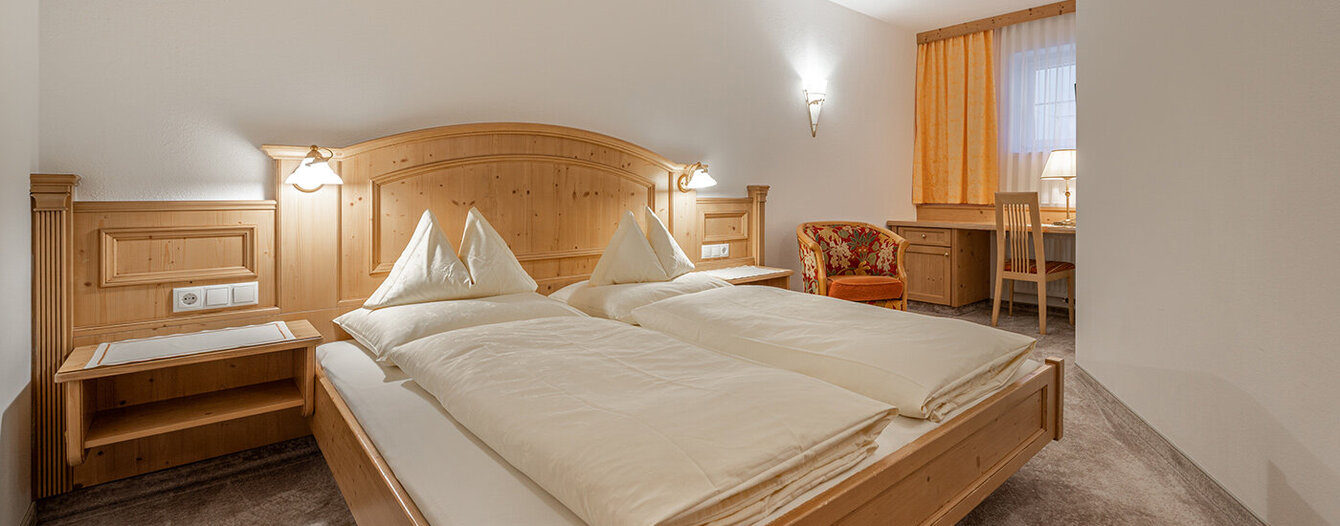 Ein gemütliches Apartmentzimmer mit einem hölzernen Doppelbett, zwei Nachttischen, weißem Bettzeug, Teppichboden, einem geblümten Sessel, einem Schreibtisch mit Stuhl und einem Fenster mit gelben Vorhängen.