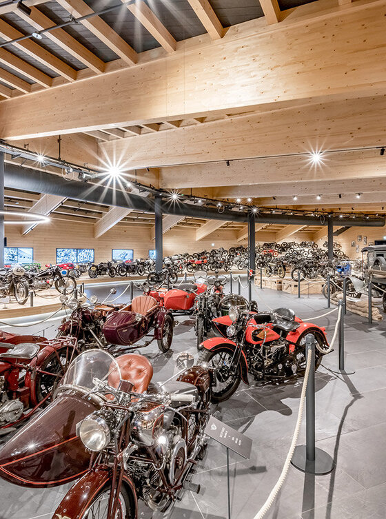 In einer geräumigen, modernen Museumsgalerie werden zahlreiche Oldtimer-Motorräder und Seitenwagen ausgestellt, wobei Holzdecken und Scheinwerfer jedes Exponat hervorheben; historische Fotos säumen die Wände im Hintergrund.