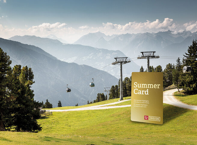 Eine sonnige Berglandschaft mit grünen Grashängen, Kiefern, Seilbahnen und einer großen Grafik der goldenen Summer Card im Vordergrund. Im Hintergrund sind dunstige, ferne Berge unter blauem Himmel zu sehen.