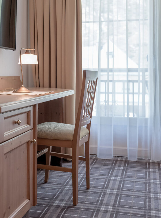 Ein kleiner Holztisch mit einem Stuhl steht an einem Fenster mit durchsichtigen Vorhängen. Auf dem Schreibtisch befinden sich eine Tischlampe, ein Notizblock und ein Stift. Ein Flachbildfernseher ist an der Wand über dem Schreibtisch angebracht.