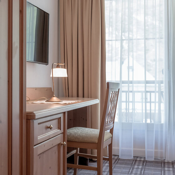 Ein kleiner Holztisch mit einem Stuhl steht an einem Fenster mit durchsichtigen Vorhängen. Auf dem Schreibtisch befinden sich eine Tischlampe, ein Notizblock und ein Stift. Ein Flachbildfernseher ist an der Wand über dem Schreibtisch angebracht.