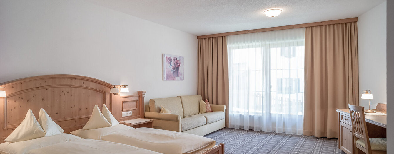 Ein helles Apartmentzimmer mit einem Doppelbett, beigem Bettzeug, einem Kopfteil aus Holz, einem Sofa, einem Schreibtisch mit Stuhl und großen Fenstern mit beigen Vorhängen, die das Tageslicht hereinlassen.