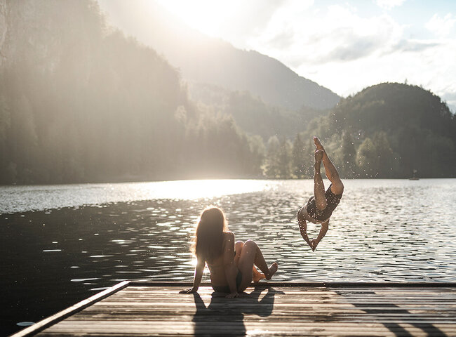 Eine Person sitzt auf einem Steg, während eine andere in den See springt, umgeben von Bergen im Sonnenlicht.