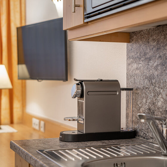 Eine moderne Küchentheke mit einer Kaffeemaschine neben einer Edelstahlspüle; im Hintergrund sind ein Flachbildfernseher, eine Lampe und gelbe Vorhänge zu sehen.