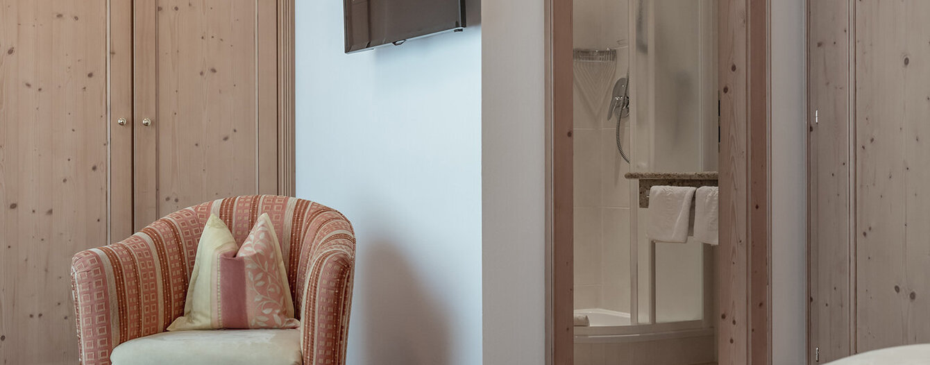 Ein gemütliches Apartmentzimmer mit einem gemusterten Sessel, einem an der Wand montierten Fernseher, Holzschränken und einer offenen Tür, die den Blick auf ein modernes Badezimmer mit Dusche und weißen Handtüchern freigibt.