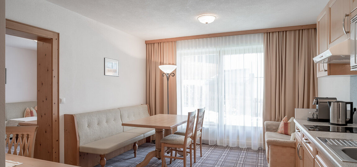 Eine helle, moderne Küche und ein Essbereich mit hellen Holzmöbeln, einer Eckbank, einem Tisch, Stühlen, einem beigen Sofa und einem großen Fenster mit durchsichtigen Vorhängen, die natürliches Licht hereinlassen.