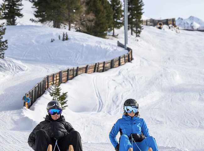 Zwei Personen mit Helmen und Winterausrüstung rodeln auf Schnee mit Skiliften und verschneiten Bergen im Hintergrund.