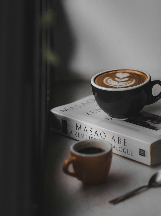 Eine schwarze Tasse Milchkaffee mit Latte Art steht auf einem Buch neben einer kleinen braunen Espressotasse und einem Löffel auf einem Tisch am Fenster.