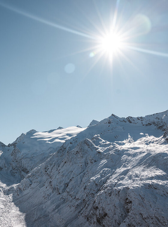 Schneebedeckte Berggipfel unter einem klaren blauen Himmel, über dem die Sonne hell scheint.