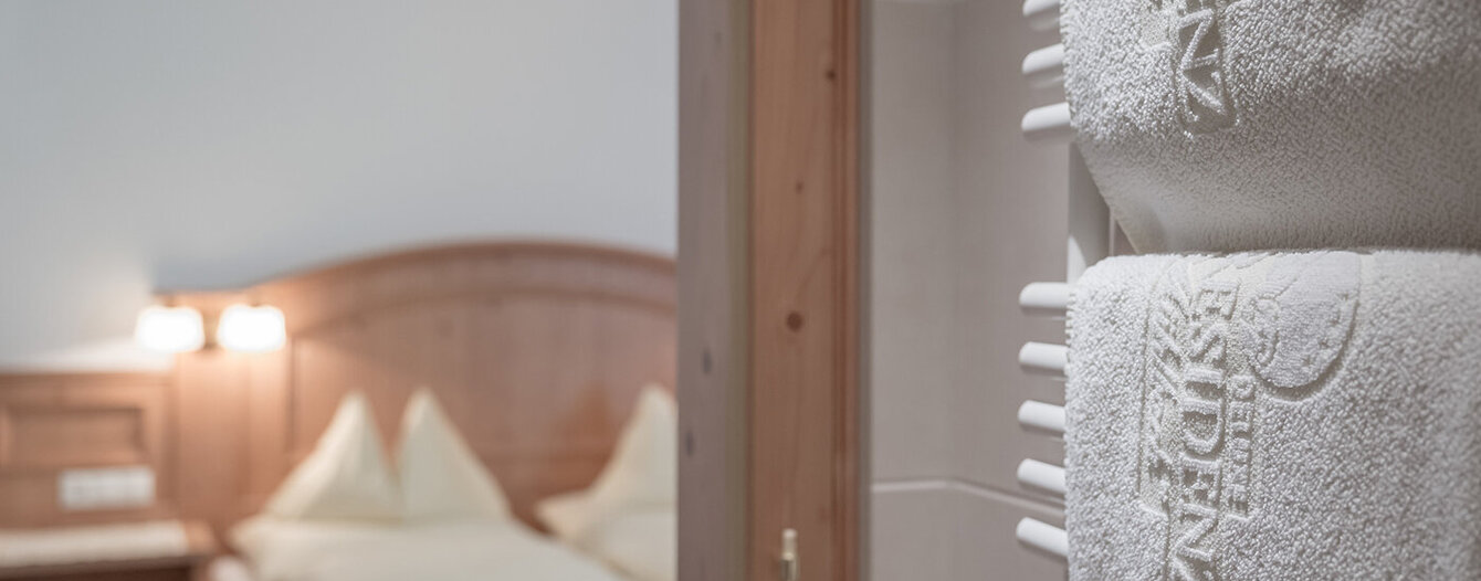 Eine Nahaufnahme von zwei weißen, bestickten Handtüchern, die auf einem Ständer hängen, mit einem unscharfen Hintergrund, der ein ordentlich gemachtes Bett und Holzmöbel in einem sanft beleuchteten Schlafzimmer zeigt.