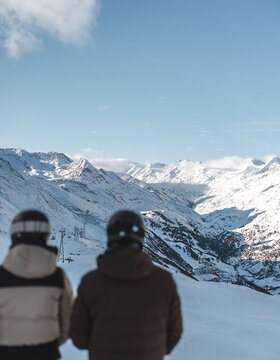Zwei Personen in Winterjacken und Helmen stehen an einem verschneiten Berghang und blicken auf eine malerische Aussicht auf schneebedeckte Gipfel unter einem klaren blauen Himmel.