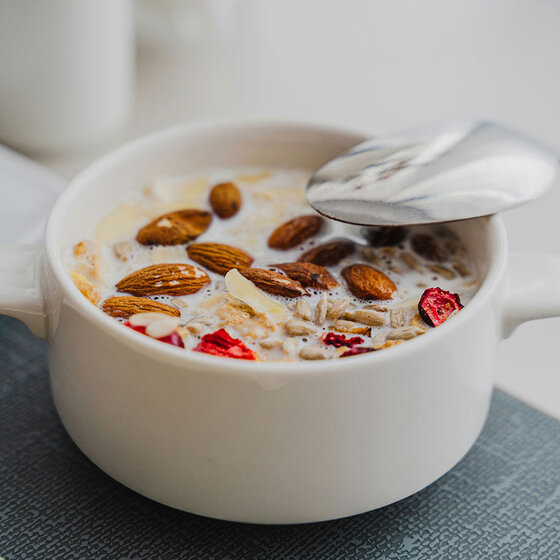 Eine weiße Schüssel, gefüllt mit Milch, Mandeln, Haferflocken, Rosinen und getrockneten roten Beeren, steht auf einem grauen Platzdeckchen. In der Schale befindet sich ein Löffel.