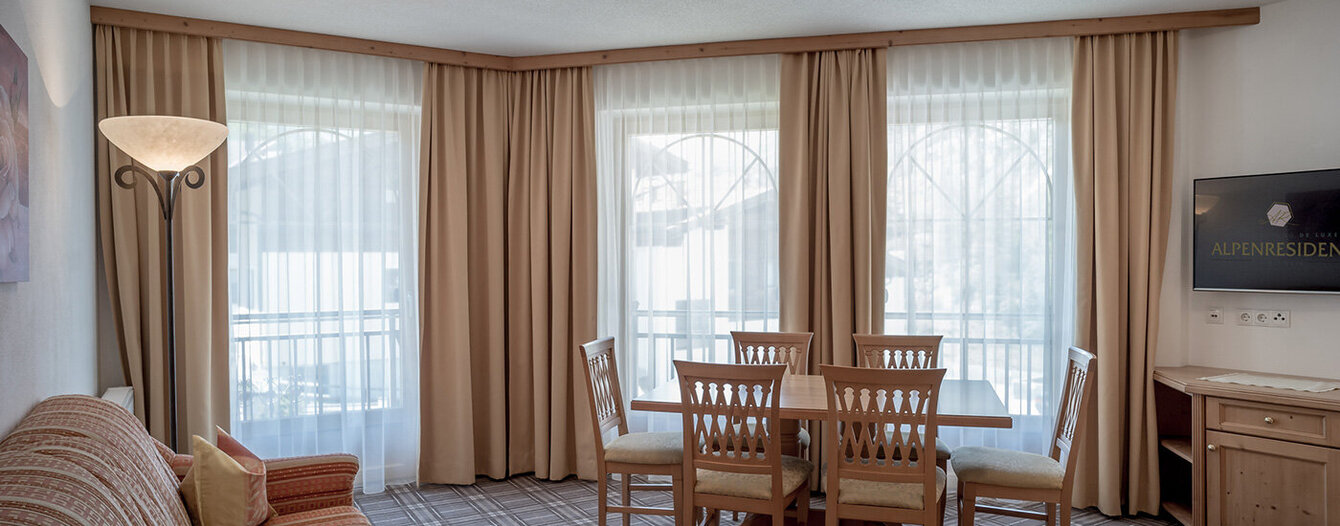 Ein heller Wohn- und Essbereich mit großen Fenstern, beigen Vorhängen, einem gemusterten Sofa, einem hölzernen Esstisch mit sechs Stühlen und einem an der Wand montierten Fernseher auf der rechten Seite. Der Raum ist sanft beleuchtet und mit einem karierten Teppichboden ausgelegt.