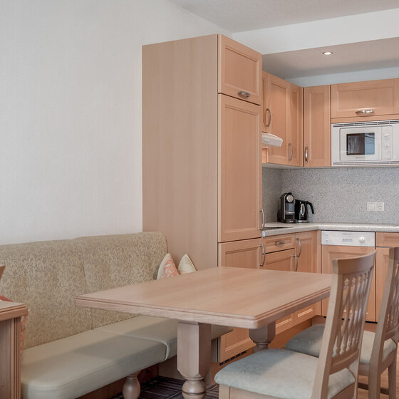 Eine kleine Küche mit hellen Holzschränken, Einbaugeräten, einer Eckbank mit Kissen und einem hölzernen Esstisch mit Stühlen, die einen gemütlichen Essbereich schaffen.