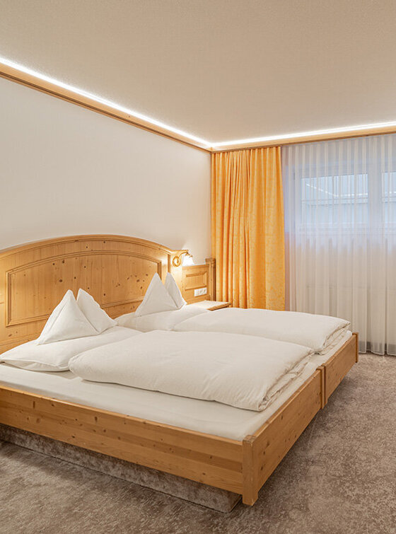 Ein gemütliches Apartmentzimmer mit Doppelbett, Holzmöbeln, Schreibtisch mit Stuhl, Wandfernseher, Kochnische mit Mikrowelle und Kaffeemaschine und einem großen Fenster mit gelben Vorhängen, durch das Tageslicht fällt.