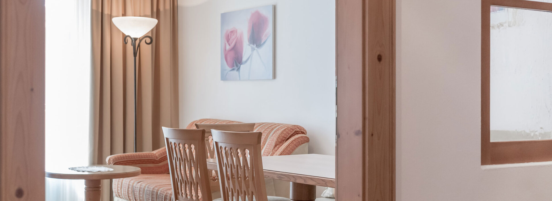 Ein gemütlicher Wohnbereich mit einem beigen Sofa, einem Esstisch und Stühlen aus Holz, einer Stehlampe und einem Gemälde mit rosa Rosen an der Wand. Licht fällt durch ein Fenster mit beigen Vorhängen ein.