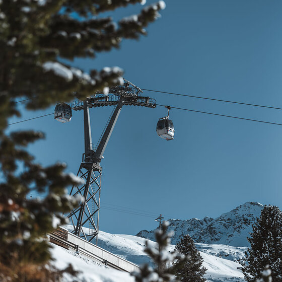 Ein Skilift mit zwei Gondeln fährt an einem klaren, sonnigen Tag über verschneite Berghänge, umrahmt von schneebedeckten Tannen, mit fernen Gipfeln im Hintergrund.
