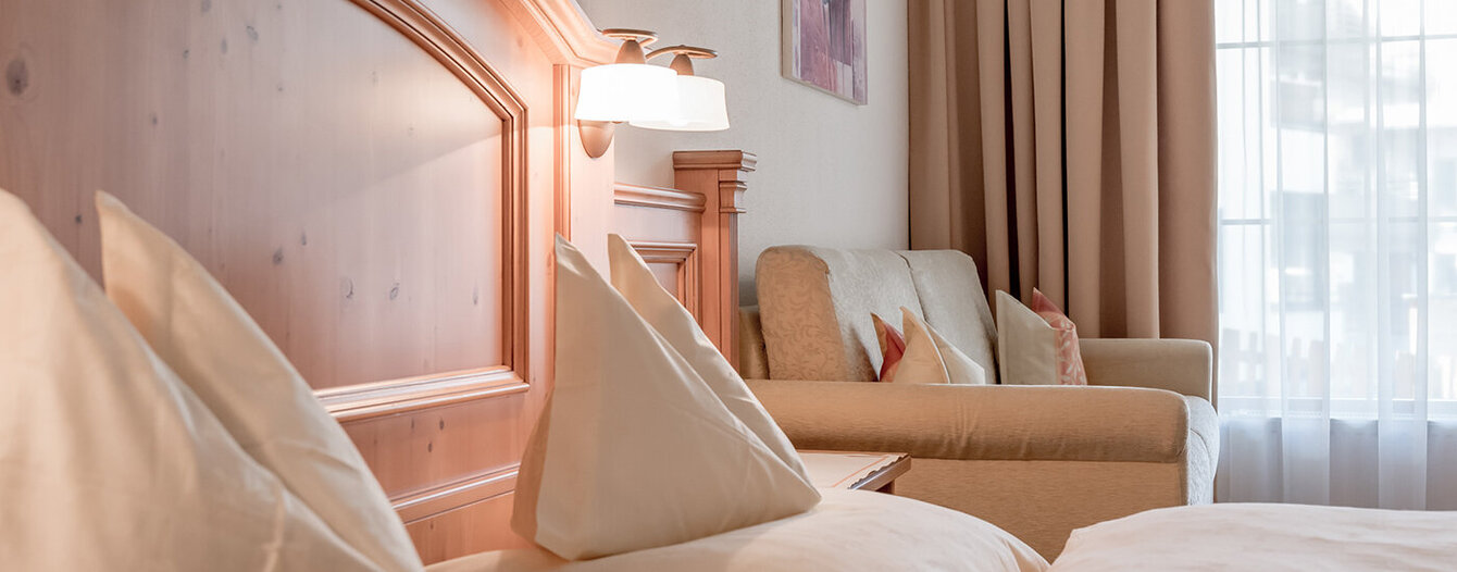 Ein gemütliches Hotelzimmer mit ordentlichen Betten mit weißer Bettwäsche, einem hellen Holzkopfteil, einem beigen Sofa mit Kissen, Wandkunst, warmer Beleuchtung und großen Vorhängen an einem hellen Fenster.