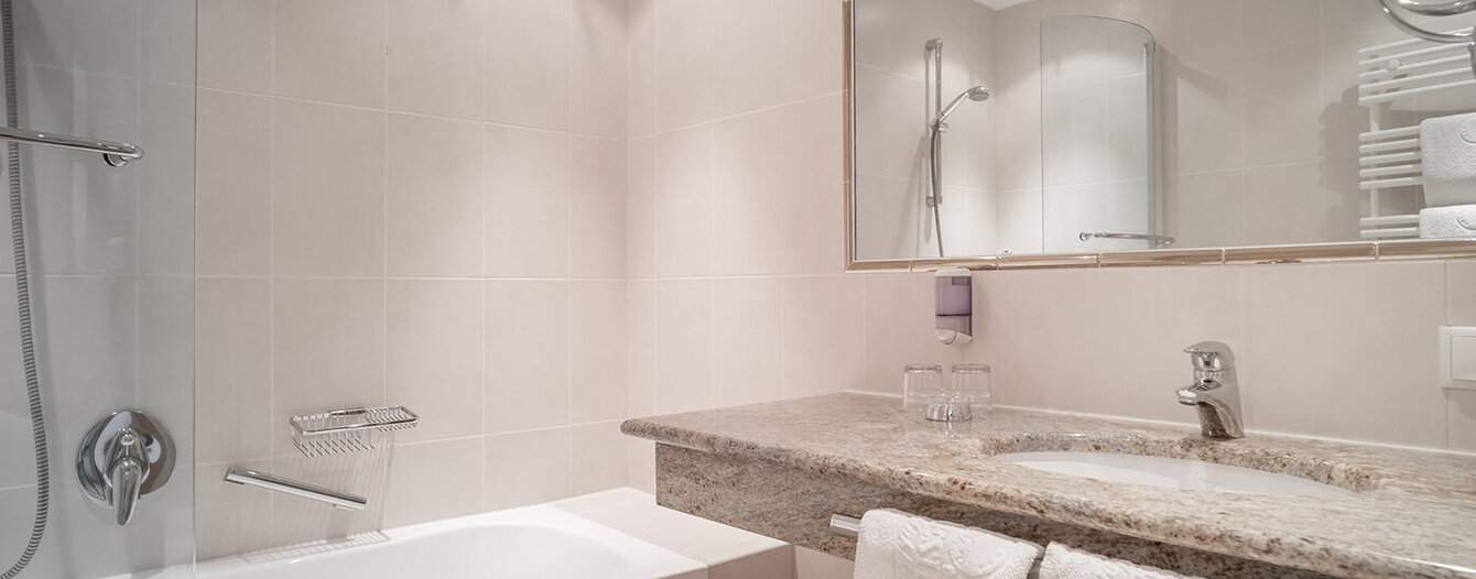 Modernes Badezimmer mit beigen Fliesen, einer Badewannen-Dusch-Kombination, einem großen Spiegel über einer Granitarbeitsplatte und weißen Handtüchern, die auf einem Regal und einer Arbeitsplatte hängen. Der Raum ist sauber und gut beleuchtet.