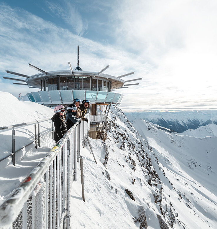 Menschen stehen auf einer schneebedeckten Aussichtsplattform in der Nähe eines modernen Rundbaus und blicken auf das Alpenpanorama unter einem teilweise bewölkten Himmel.