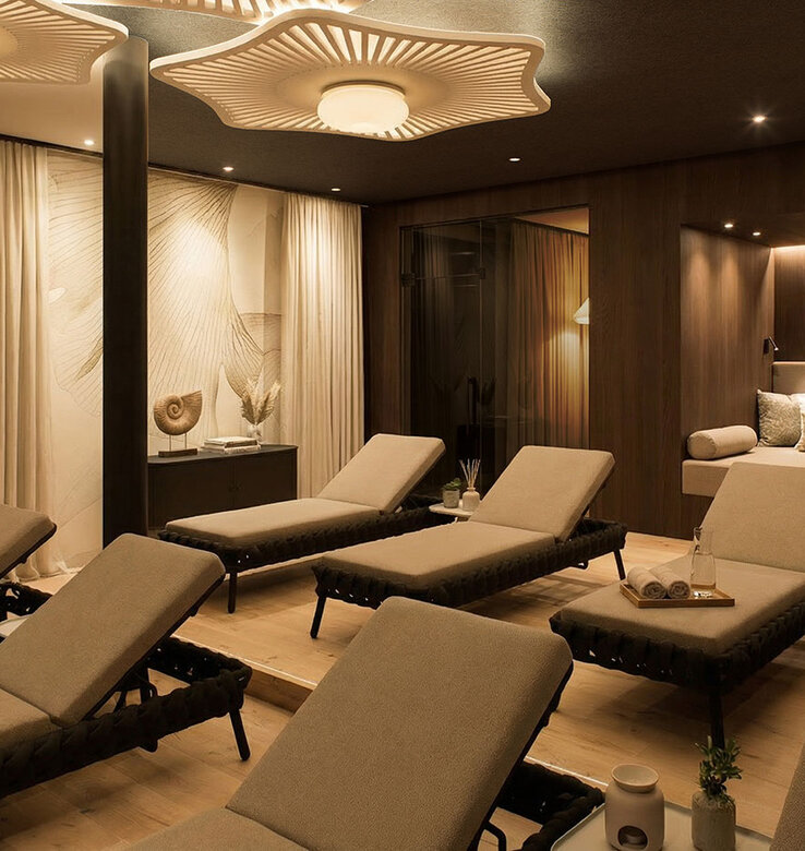 Ein luxuriöser Spa-Entspannungsraum mit mehreren Wellnessliegen, die jeweils mit einem aufgerollten Handtuch und einem Tablett ausgestattet sind, warmer Umgebungsbeleuchtung, moderner Einrichtung, gemütlichen Sitzecken und eleganten Holz- und Stoffakzenten.