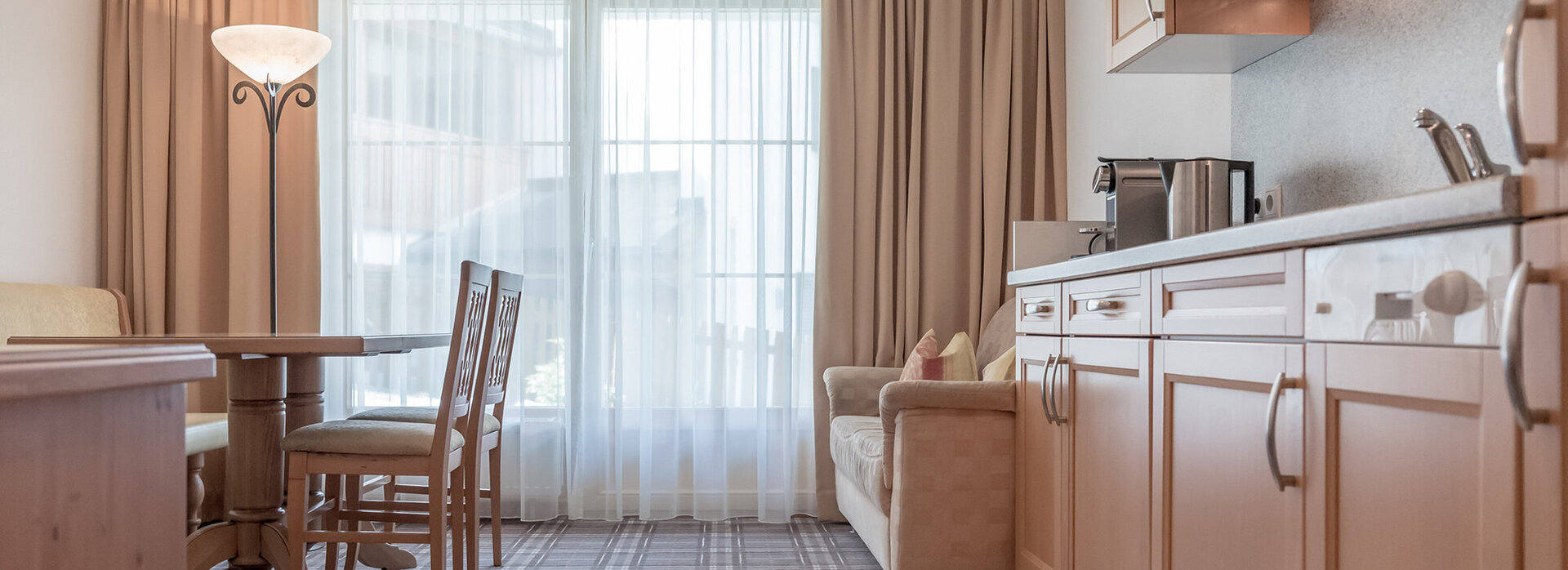 Helle Küche und Essbereich mit Holzschränken, Tisch und Stühlen, einem beigen Sofa, einer Stehlampe und einem großen Fenster mit durchsichtigen Vorhängen, die natürliches Licht hereinlassen. Der Boden ist eine Mischung aus Fliesen und Teppich.