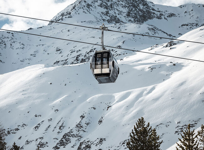 Eine Seilbahn, die an einem Seil aufgehängt ist, fährt über verschneite Berghänge, unter denen sich Kiefern befinden, mit einem schroffen, schneebedeckten Gipfel im Hintergrund und einem teilweise bewölkten Himmel.