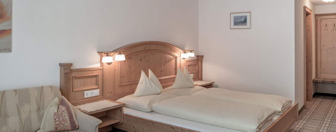 Ein gemütliches Hotelzimmer mit einem Doppelbett aus Holz, zwei Nachttischlampen, einem beigen Sofa, einem kleinen Tisch mit einem Tuch und Teppichboden. Die Wände sind weiß mit einem gerahmten Bild und einer Leuchte an der Decke.