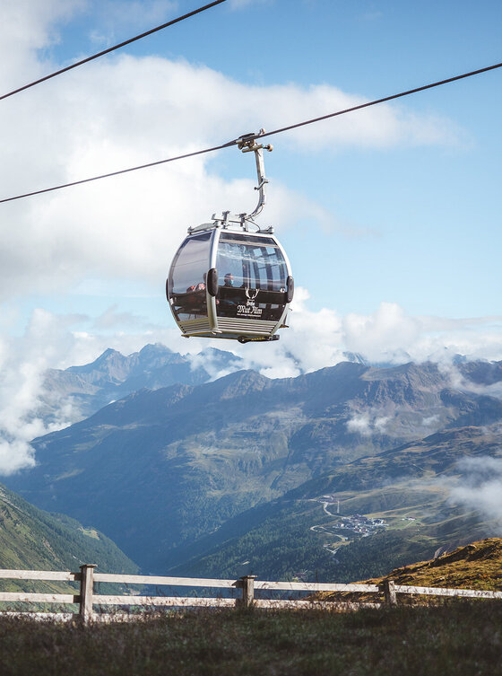 Eine Gondelbahn fährt an einem Seil hoch über eine Berglandschaft mit sanften grünen Hügeln, gewundenen Straßen und Wolken am blauen Himmel.