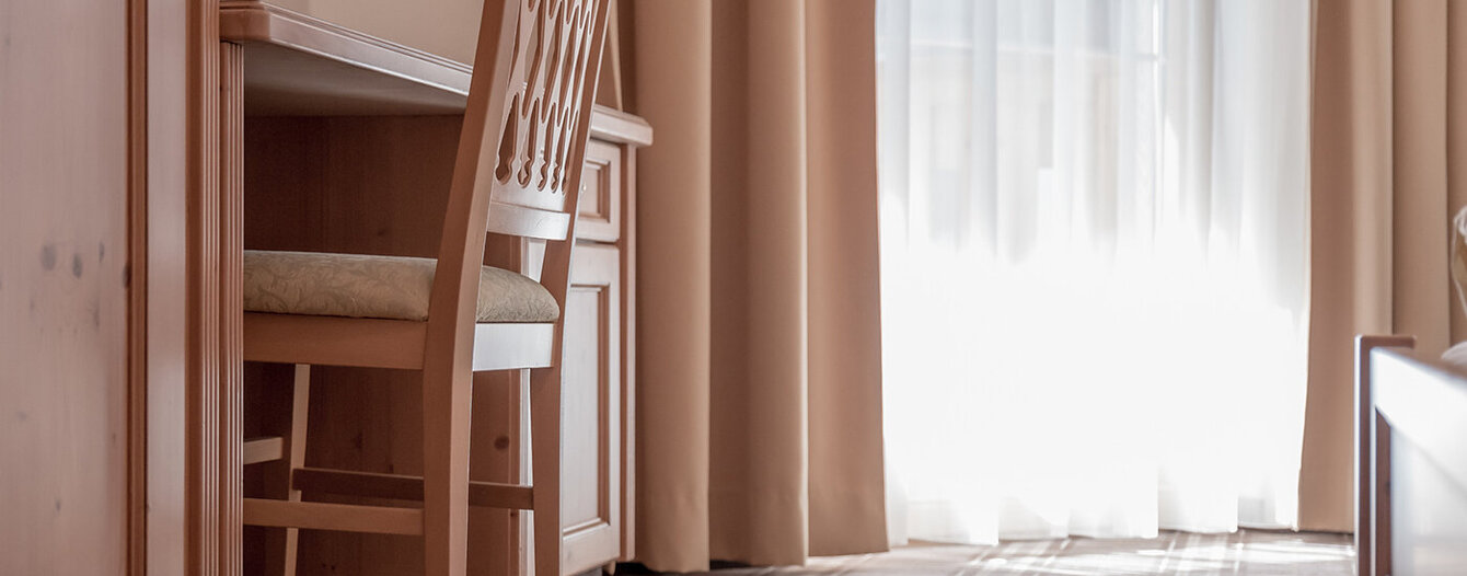 Ein Holzstuhl steht unter einem passenden Holzschreibtisch in einem sanft beleuchteten Raum mit beigen Vorhängen und einem Fenster, das natürliches Licht hereinlässt. Der Teppich hat ein dezentes Karomuster.