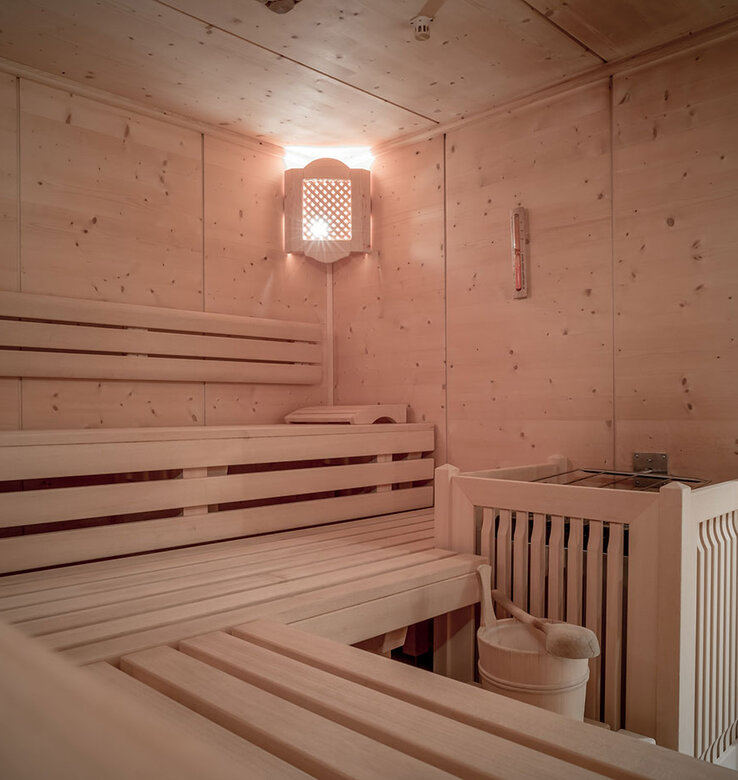 Eine gemütliche Holzsauna mit gestuften Sitzbänken, einem Eimer und einer Schöpfkelle auf der unteren Bank, einem Ofen mit Steinen in der Ecke und warmem Licht, das den Raum beleuchtet.