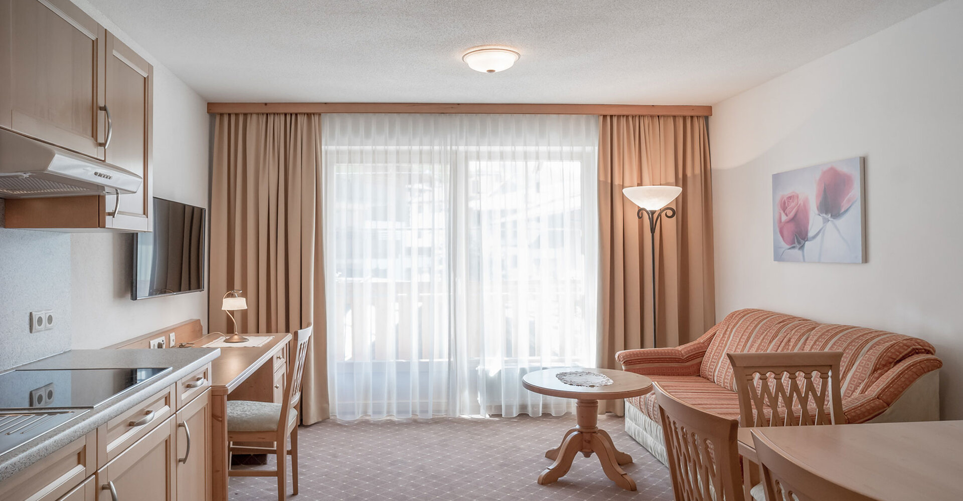 Ein heller, aufgeräumter Wohnraum mit einem Sofa, einem runden Couchtisch, Holzstühlen und einem Schreibtisch an einem Fenster mit transparenten Vorhängen. Auf der linken Seite befindet sich eine Küchenzeile, und der Raum ist in sanften, neutralen Tönen gehalten.