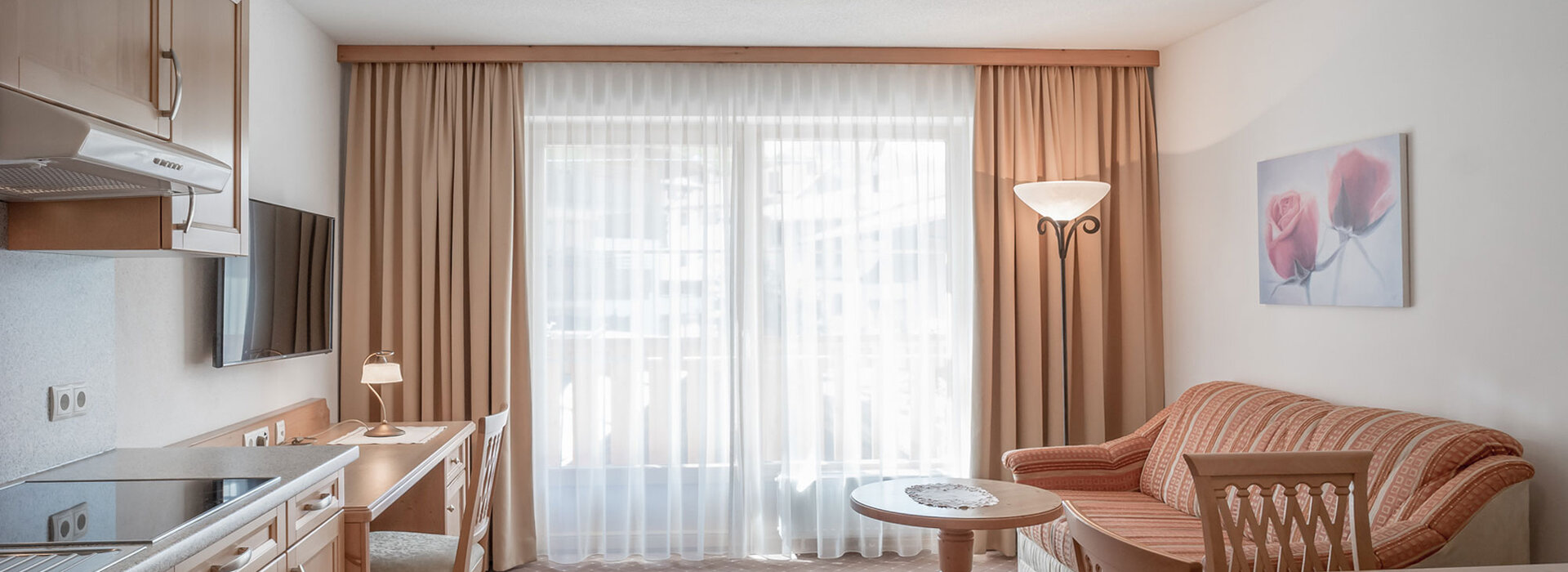 Ein heller, aufgeräumter Wohnraum mit einem Sofa, einem runden Couchtisch, Holzstühlen und einem Schreibtisch an einem Fenster mit transparenten Vorhängen. Auf der linken Seite befindet sich eine Küchenzeile, und der Raum ist in sanften, neutralen Tönen gehalten.