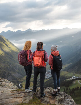 Drei Personen mit Rucksäcken stehen auf einem Felsvorsprung mit Blick auf ein malerisches Gebirgstal, in dem die Sonnenstrahlen durch die Wolken brechen und die Landschaft unter ihnen beleuchten.