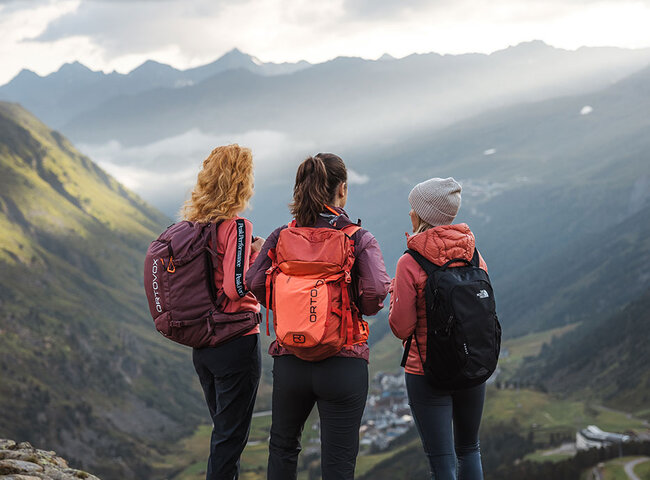 Drei Personen mit Rucksäcken stehen auf einem Felsvorsprung mit Blick auf ein malerisches Gebirgstal, in dem die Sonnenstrahlen durch die Wolken brechen und die Landschaft unter ihnen beleuchten.