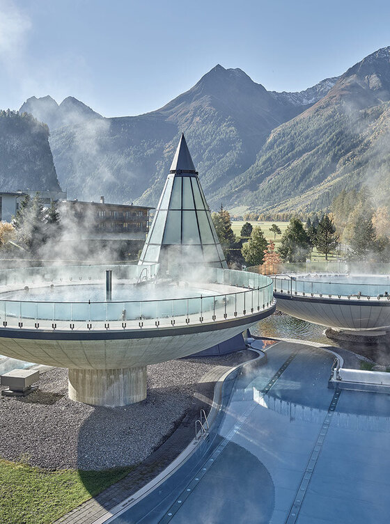 Zwei große, futuristisch anmutende Thermalbecken im Freien geben in einer Berglandschaft Dampf ab, in deren Nähe sich eine gläserne Pyramide befindet, während im Hintergrund bewaldete Hügel und Gipfel unter einem klaren Himmel zu sehen sind. Gezeigt wird das Aqua Dome in Längenfeld.