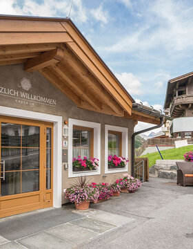 Eingang eines gemütlichen Alpengasthofs mit Blumenkästen, einer Holztür und einem Schild mit der Aufschrift Herzlich Willkommen. Im Hintergrund sind Sitzgelegenheiten im Freien und ein weiteres Haus mit Balkonen und Blumen zu sehen.