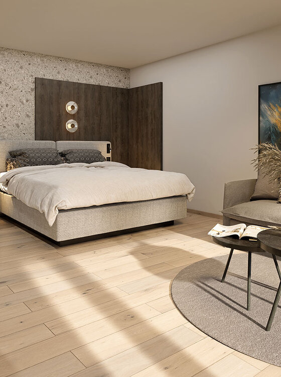 Modernes Schlafzimmer mit hellem Holzboden, einem großen Bett, einem grauen Sofa mit Couchtisch, einem runden Teppich, Wandkunst und dekorativer Beleuchtung. Der Raum wirkt offen und stilvoll mit neutralen Tönen und minimalem Dekor.