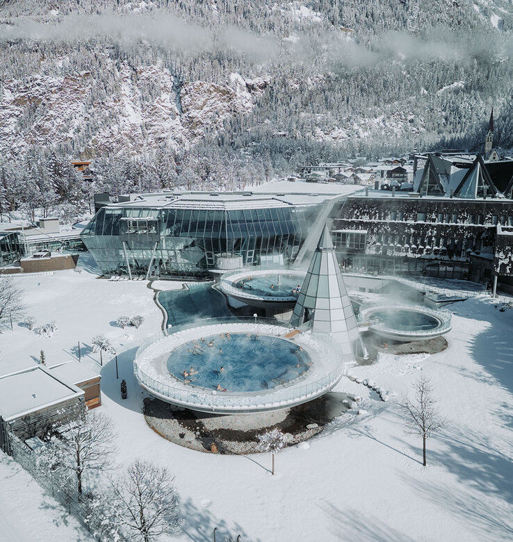 Luftaufnahme vom Aqua Dome mit Whirlpools im Freien mit aufsteigendem Dampf, umgeben von Schnee und modernen Glasgebäuden in einer verschneiten Berglandschaft.