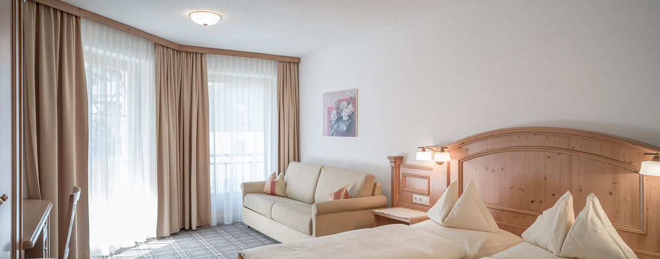 Ein helles Apartmentzimmer mit sauber gemachtem Doppelbett, einem beigen Sofa, einem Schreibtisch und einem Stuhl, großen Fenstern mit hellen Vorhängen und einem gerahmten Bild an der weißen Wand. Das Zimmer ist sanft beleuchtet und hat einen Teppichboden.