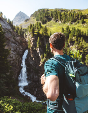 Ein Mann mit Rucksack steht an einem hellen, sonnigen Tag vor einem Wasserfall und üppigen Bergen.