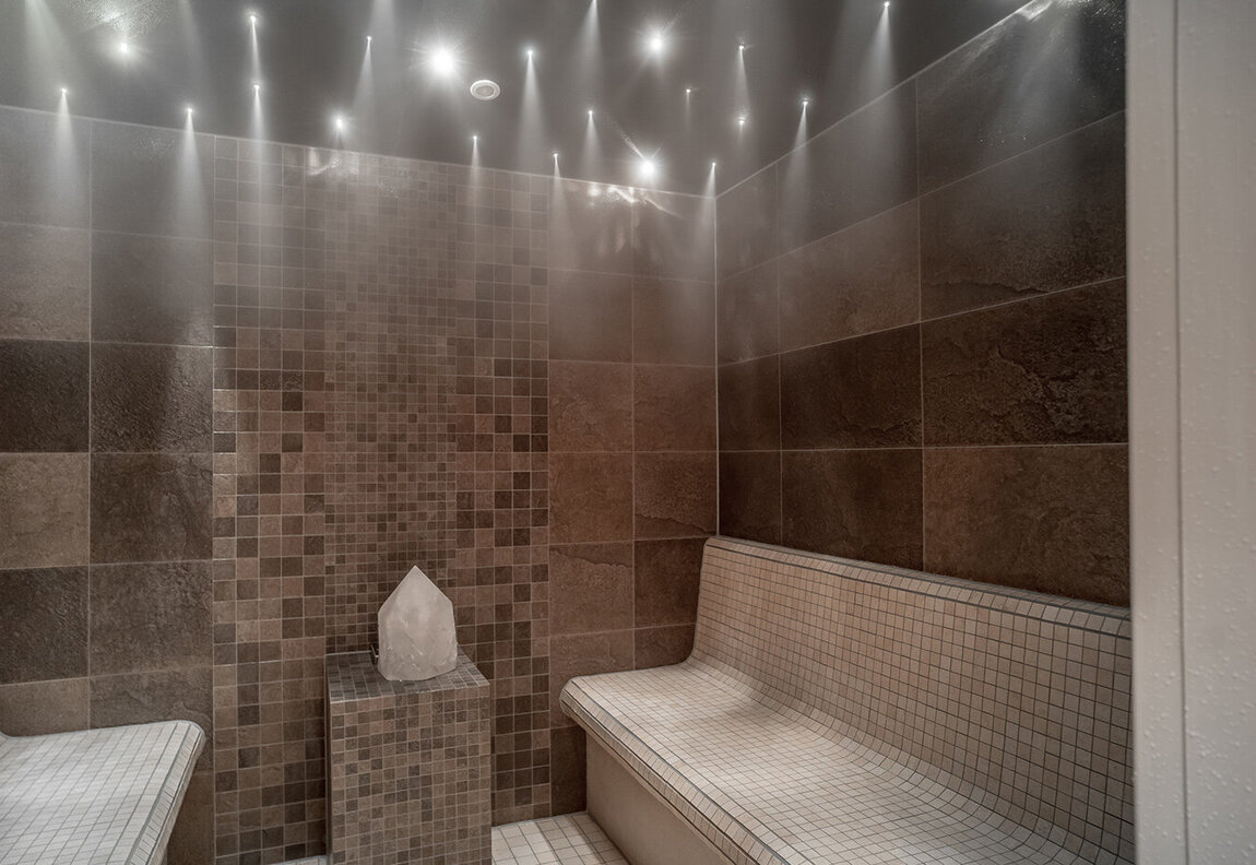 Ein modernes Dampfbad mit gefliesten Wänden und Bänken, Mosaikfliesen als Akzente, einem Kristall in der Mitte des Raumes auf einem Sockel und sternförmigen Deckenleuchten, die eine entspannende Atmosphäre schaffen.