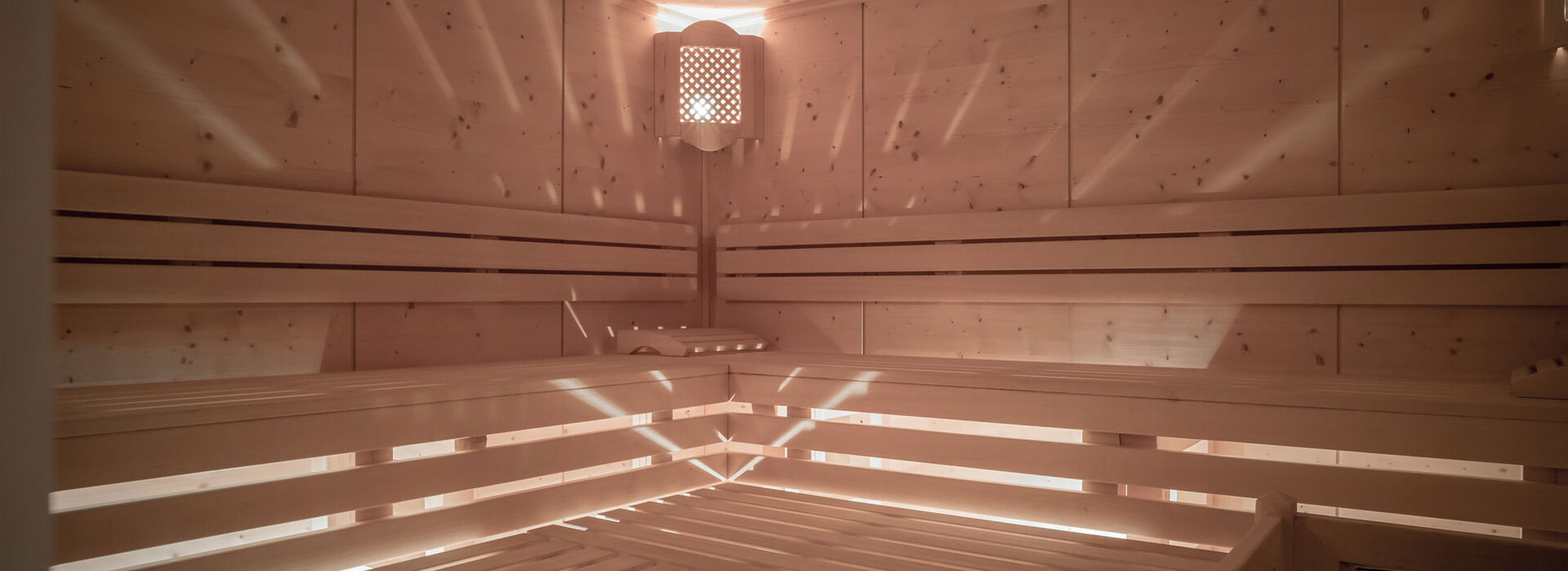 Eine geräumige Holzsauna mit abgestuften Bänken, einem Eimer und einer Kelle, einem Wandofen und sanftem natürlichem Licht, das durch Lamellen strömt und Muster an den Wänden und Bänken erzeugt.