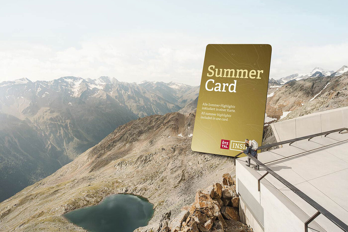 Eine große und goldene Grafik mit der Aufschrift "Summer Card" steht auf einer Bergterrasse mit Blick auf eine felsige Landschaft mit einem kleinen See und schneebedeckten Gipfeln unter einem bewölkten Himmel.