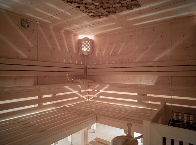 Das Innere einer Holzsauna mit gestuften Bänken, sanftem Licht, das durch die Lattenwände und die Decke fällt, einer Wanduhr, einem Eimer mit Schöpfkelle und einem Ofen in der Ecke.