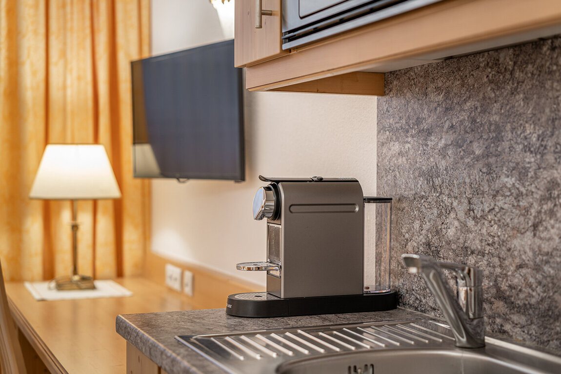Eine moderne Küchenarbeitsplatte mit einer Kaffeemaschine neben einem Waschbecken. Im Hintergrund befinden sich ein Flachbildfernseher und eine Lampe auf einem Holztisch mit gelben Vorhängen dahinter.
